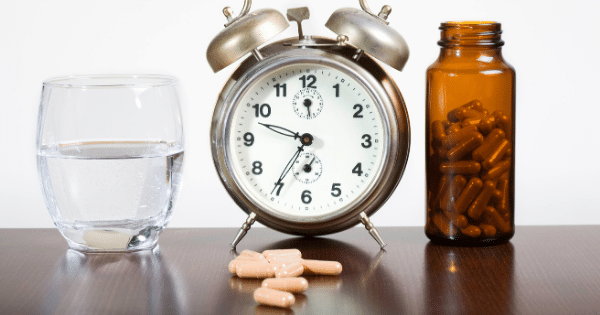A traditional silver twin-bell alarm clock sits in the center of a dark wooden table, flanked by a glass of water on the left and an amber pill bottle filled with brown capsules on the right. A small pile of matching loose capsules is scattered on the table directly in front of the clock.