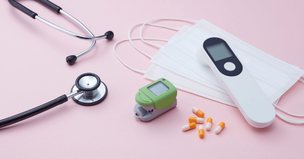A collection of medical equipment arranged neatly on a smooth pink surface. It includes a black stethoscope on the left, a stack of white disposable medical masks, a white infrared forehead thermometer, a green fingertip pulse oximeter, and a small scatter of orange and white capsules.