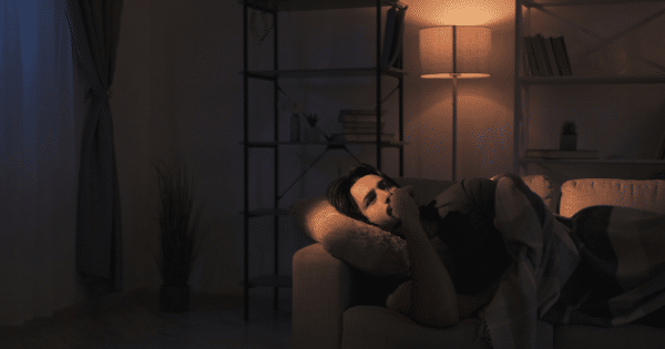 A young man lies on a couch in a dimly lit living room at night, tucked under a blanket. He is awake, looking up thoughtfully with his hand near his mouth. In the background, a tall bookshelf stands next to a lit floor lamp casting a warm glow.