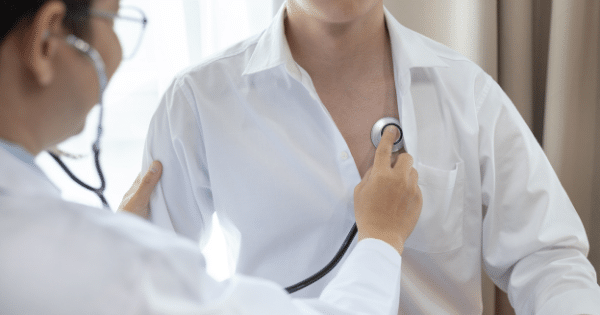 A close-up view of a doctor wearing glasses and a white coat using a stethoscope to check the heartbeat of a male patient. The patient is wearing a white button-down shirt that is slightly open at the chest where the stethoscope chest piece is placed.