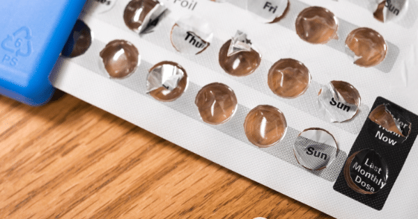 A close-up, angled shot of an opened birth control pill blister pack resting on a wooden surface. Several of the foil compartments have been broken open, revealing empty slots, with days of the week like "Thu" and "Sun" printed next to the bubbles. A blue plastic case edge is visible on the left.