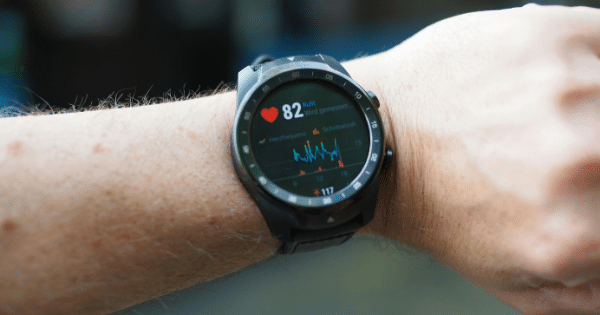 A close-up view of a person’s wrist wearing a black smartwatch that displays health tracking metrics. The screen shows a heart rate of 82 bpm with a red heart icon, alongside a real-time heart rate graph line and other German text indicators.