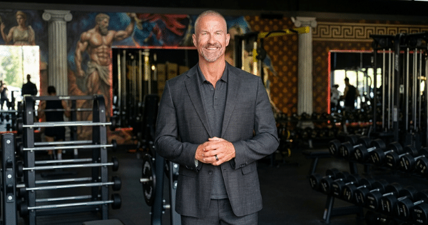 A medium shot of a smiling, fit middle-aged man with a shaved head and graying beard, standing in the center of a spacious gym. He is wearing a dark gray textured suit jacket over a matching dark polo shirt, with his hands clasped together in front of him.