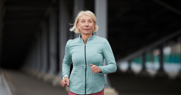 A middle-aged woman with short blonde hair is jogging directly toward the camera under a concrete bridge or overpass structures. She is wearing a light teal zip-up athletic jacket over pink pants, with a subtle, focused expression on her face.