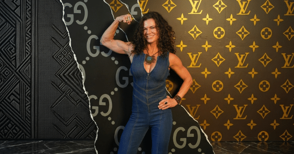 A smiling, muscular woman with dark curly hair poses against a stylized wall background. The wall has a torn effect separating a dark pattern with repeating "GG" logos on the left from a gold patterned background with repeating "LV" logos on the right. She is wearing a denim sleeveless jumpsuit with a front zipper and is flexing her right bicep.