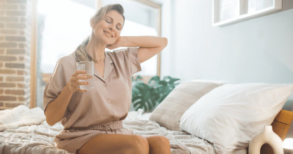 A relaxed woman sits on a bed with neutral-toned bedding, wearing silky beige pajamas. She holds a glass of water in one hand and stretches her other arm behind her head with her eyes closed and a contented smile. Sunlight streams through a window in the background near a large green houseplant.