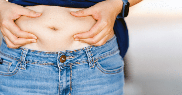 A close-up shot focusing on a person's midsection as they pinch a fold of skin and fat on their belly with both hands. The individual is wearing light-blue denim jeans and a dark blue shirt that is pulled up to expose the midriff.