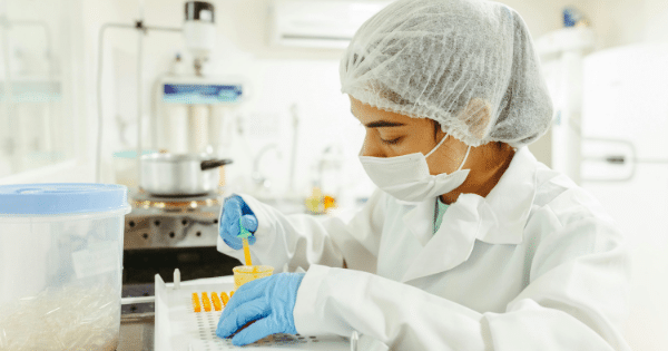 A laboratory technician wearing a white lab coat, a white hairnet, a face mask, and blue nitrile gloves sits at a workstation. She is carefully using a small pipette to dispense a yellow liquid into a multi-slot tray filled with clear capsules.