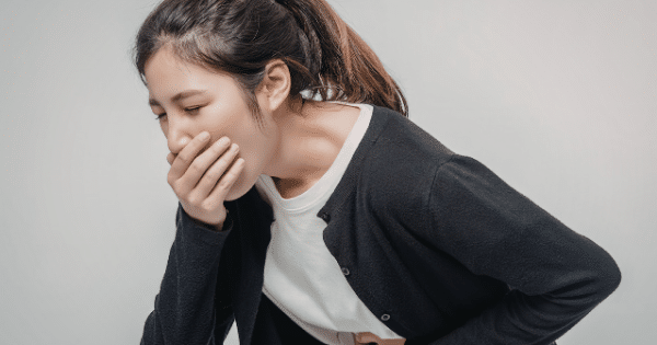 A young woman with her dark hair tied back in a ponytail is leaning forward against a plain light gray background. She has one hand over her mouth and the other pressed against her stomach, looking distressed as if experiencing nausea or an urge to vomit.