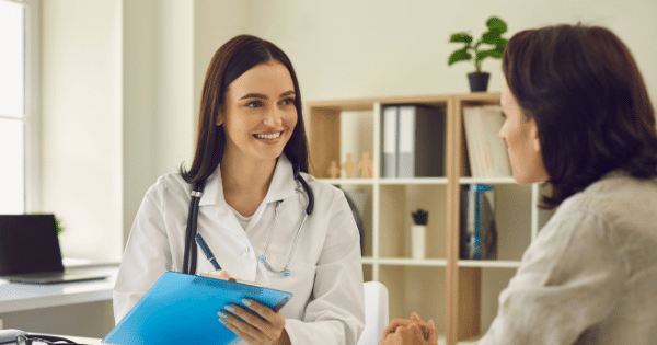 A smiling female doctor with long dark hair, wearing a white medical coat and a stethoscope around her neck, sits at a desk while consulting with a patient. The doctor holds a blue clipboard and pen, looking attentively toward the patient whose back is partially turned to the camera.