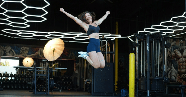 A fit woman with dark curly hair is captured mid-air as she enthusiastically jumps inside a gym with her arms raised wide in celebration. She is wearing a black sports bra, dark blue athletic shorts, and white sneakers. The gym ceiling features modern, glowing hexagonal light fixtures.