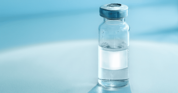 A close-up shot of a small, clear glass medical vial with a blank white label and a silver metal cap, sitting against a soft blue, backlit background.