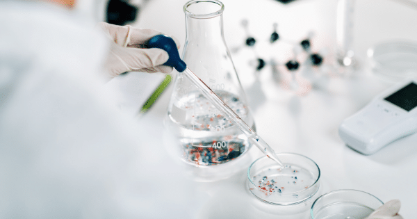 An over-the-shoulder view of a scientist in a white lab coat and gloves using a pipette with a blue bulb to drop liquid into a petri dish containing small red and blue particles.