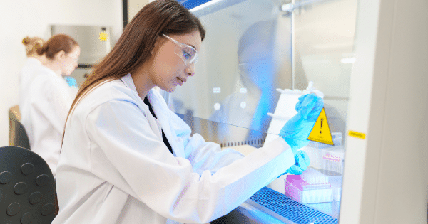 A female scientist wearing safety glasses and a lab coat working inside a sterile laboratory hood. She is wearing blue gloves and handling small tubes near a yellow biohazard warning sticker.