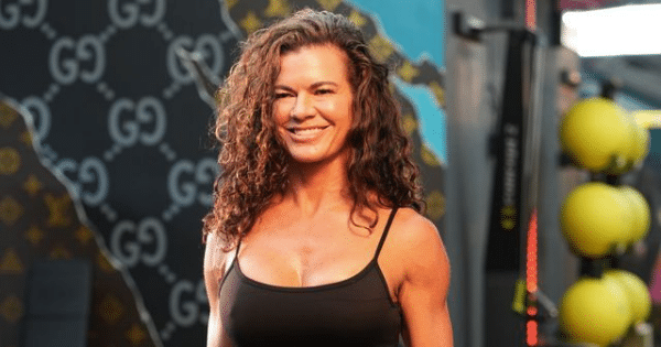 A portrait of a smiling woman with long, curly brown hair wearing a black tank top, standing in a gym setting. The background features a dark wall with a subtle pattern and a rack of yellow medicine balls.