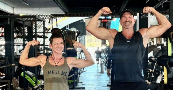 A woman in a green tank top that reads "ME VS. ME" and a man in a black tank top and baseball cap both smiling and flexing their biceps together inside a gym filled with workout equipment.