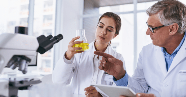 An older male scientist with glasses and a young female scientist, both wearing white laboratory coats, collaborate in a bright lab. The woman holds up an Erlenmeyer flask filled with yellow liquid for inspection while the man gestures toward it and looks at a digital tablet.