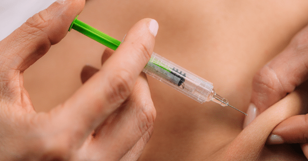 A close-up shot of a person's hands performing a subcutaneous injection into a patch of pinched skin. The clear plastic syringe features a bright green plunger and contains a small dose of medication.