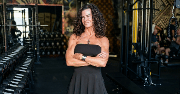 A woman with curly brown hair wearing a strapless black dress stands in a gym with her arms crossed, smiling directly at the camera. In the background, rows of dumbbells and weight machines are visible.