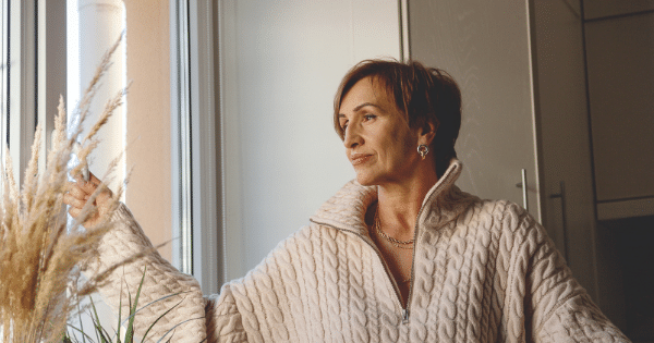 A woman with short brown hair wearing a chunky cream-colored cable-knit sweater stands indoors by a window, looking thoughtfully off to the side while gently touching a vase of dried pampas grass.
