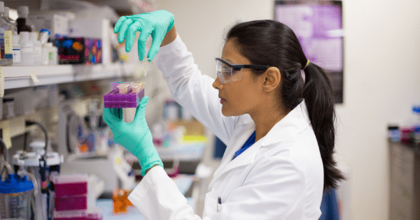 A female scientist with her hair in a ponytail, wearing safety glasses, a white lab coat, and green gloves, stands in a laboratory. She is holding a purple test tube rack filled with small vials in one hand while using her other hand to select or adjust a vial.