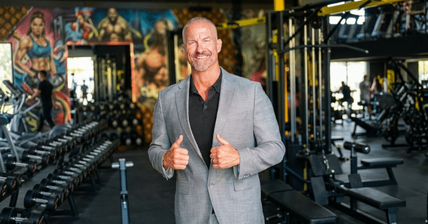 A smiling, athletic middle-aged man in a gray suit jacket and a black button-down shirt stands in the center of a spacious gym giving a double thumbs-up. The background shows rows of dumbbells, weight machines, and a colorful fitness-themed wall mural.