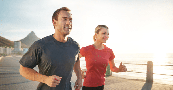 A man in a dark gray athletic shirt and a woman in a red t-shirt jog side-by-side along an oceanfront boardwalk during a bright sunset. In the distant background, a mountain peak and coastal buildings are visible under a clear sky.