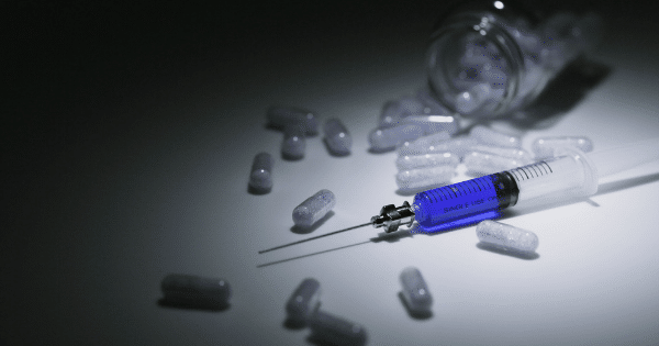 A medical syringe filled with vibrant blue fluid lies diagonally on a white surface under a spotlight. Scattered around the syringe are numerous clear capsules filled with white granular powder, with an overturned glass jar visible in the dimly lit background.