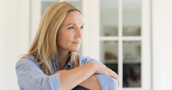 A medium shot of a blonde woman with green eyes, wearing a light blue button-up shirt. She is sitting indoors with her arms crossed over her knees, looking thoughtfully out of frame to the right.