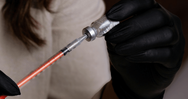 A close-up view of hands wearing black nitrile gloves. One hand holds a small glass vial upside down while the other hand inserts a thin needle to draw out an orange liquid into a syringe.