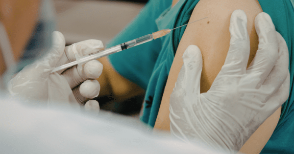 A close-up shot of a healthcare worker in white medical gloves administering a vaccine or injection using a clear syringe into the upper shoulder muscle of a patient wearing a green shirt.
