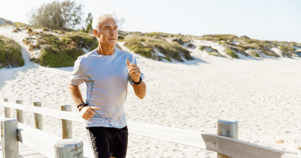 A fit, senior man with grey hair jogging along a wooden beach boardwalk on a sunny day. He is wearing a grey athletic t-shirt and black shorts, with sandy dunes visible in the background.