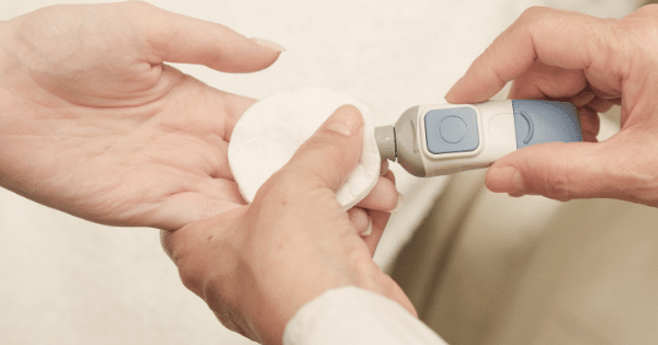 A close-up view of a person's hand being held by a healthcare provider who is using a grey and blue lancing device to perform a finger-prick blood test, while holding a white cotton pad underneath the finger.