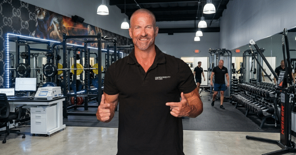 A smiling, muscular, bald man in a black polo shirt stands in the center of a well-lit, modern gym. The background shows weightlifting cages, racks of dumbbells, mirrors, and a computer desk setup on the left.