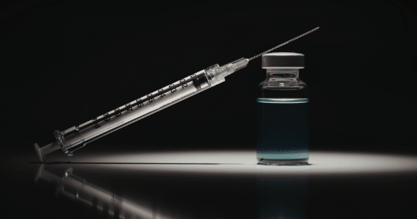 A clear medical syringe rests diagonally against a small glass vial filled with blue liquid, highlighted by a subtle spotlight on a dark, reflective surface.