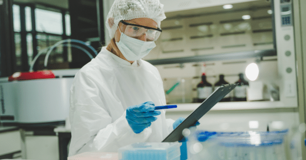 A laboratory scientist wearing a white protective gown, face mask, hairnet, and safety goggles looks down to write on a clipboard with a blue pen.