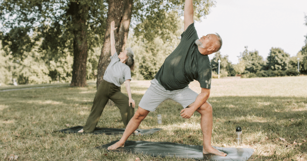An older man and woman practice yoga outside on mats in a grassy park, holding an extended side angle pose under the shade of large trees.