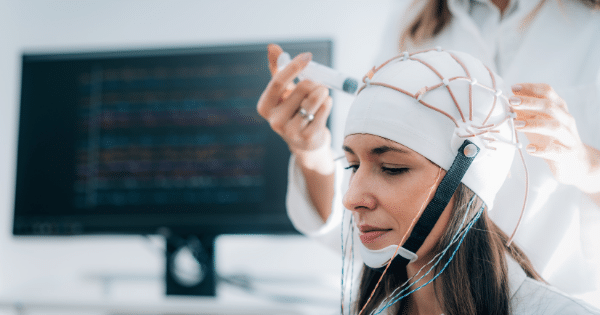 A woman wears a white EEG cap with attached wires while a medical professional in a white coat stands behind her, using a syringe to apply conductive gel to the sensors.