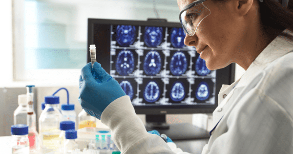 A female scientist wearing protective eyewear and blue gloves closely examines a small laboratory vial, with a computer monitor displaying brain scan images in the background.