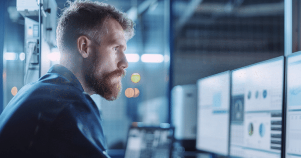 A side-profile view of a bearded looking intently at multiple computer monitors displaying analytical charts and data inside a dimly lit lab.