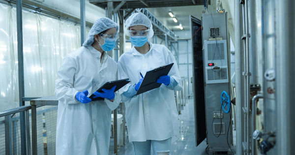 Two quality control workers in an industrial facility wearing white lab coats, face masks, hairnets, and blue gloves walk down a corridor while reviewing data on a tablet and a clipboard.