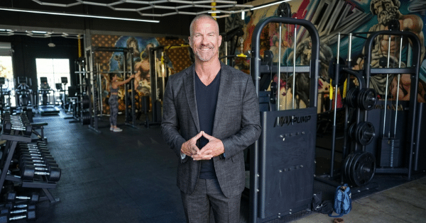 A smiling, fit man in a dark textured suit and black v-neck shirt stands in the center of a spacious gym with his hands clasped together, surrounded by workout equipment, weight racks, and colorful wall murals.