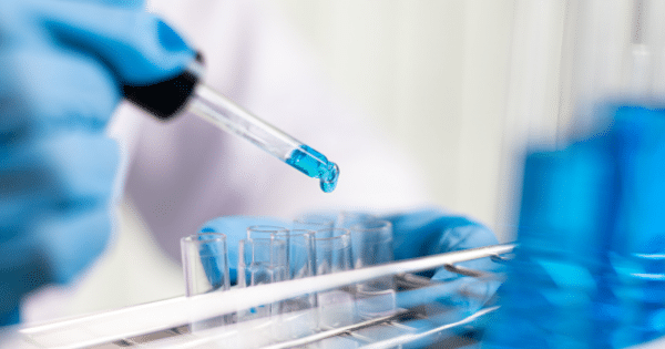 Close-up of a hand in a blue lab glove using a pipette to drop a bright blue liquid into clear test tubes sitting in a laboratory rack.
