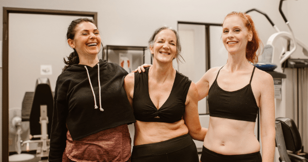 Three women of diverse ages smiling and laughing together while standing arm-in-arm inside a gym, wearing athletic workout clothes.