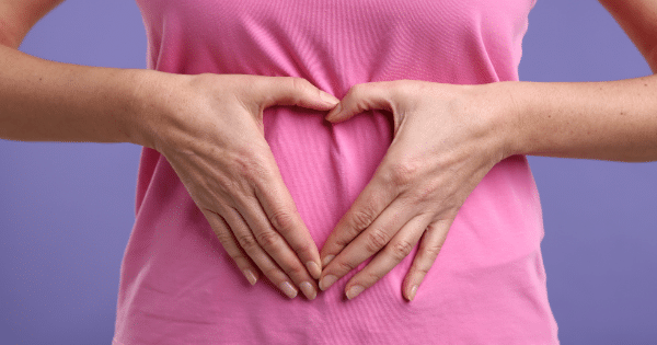 Close-up of a person wearing a pink shirt against a solid purple background, placing their hands over their abdomen to form a heart shape.