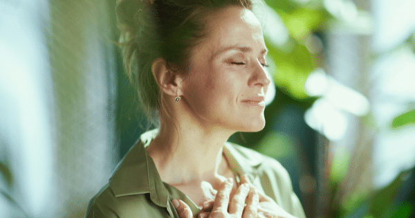 A woman with her eyes closed smiling peacefully with her hands pressed against her chest, surrounded by soft morning light and blurred green foliage.