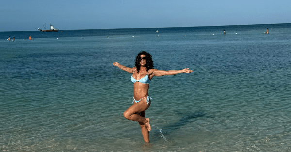 A fit woman with curly dark hair wearing a light blue bikini, posing on one leg with her arms outstretched in the calm, clear ocean water under a blue sky.