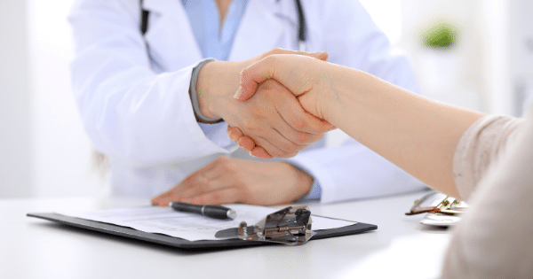 Close-up of a doctor in a white lab coat shaking hands with a patient over a medical consultation desk that has a clipboard and pen on it.