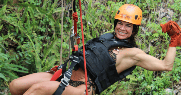 A smiling, muscular woman wearing an orange safety helmet, a life jacket, and a harness flexes her bicep while suspended on a zipline or rappelling system against a lush, green background.