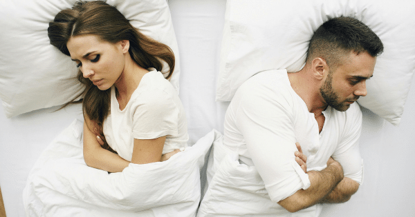 Overhead view of a man and woman lying in bed on white pillows, turned completely away from each other with arms crossed, reflecting relationship tension.